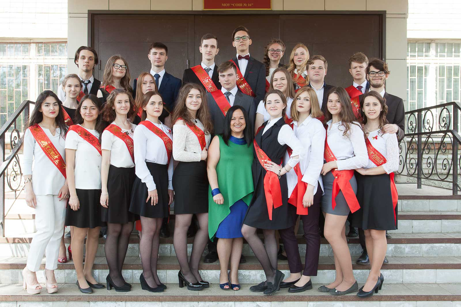 Общая фотография выпускного класса с классным руководителем в зеленом платье на ступеньках школьного крыльца.