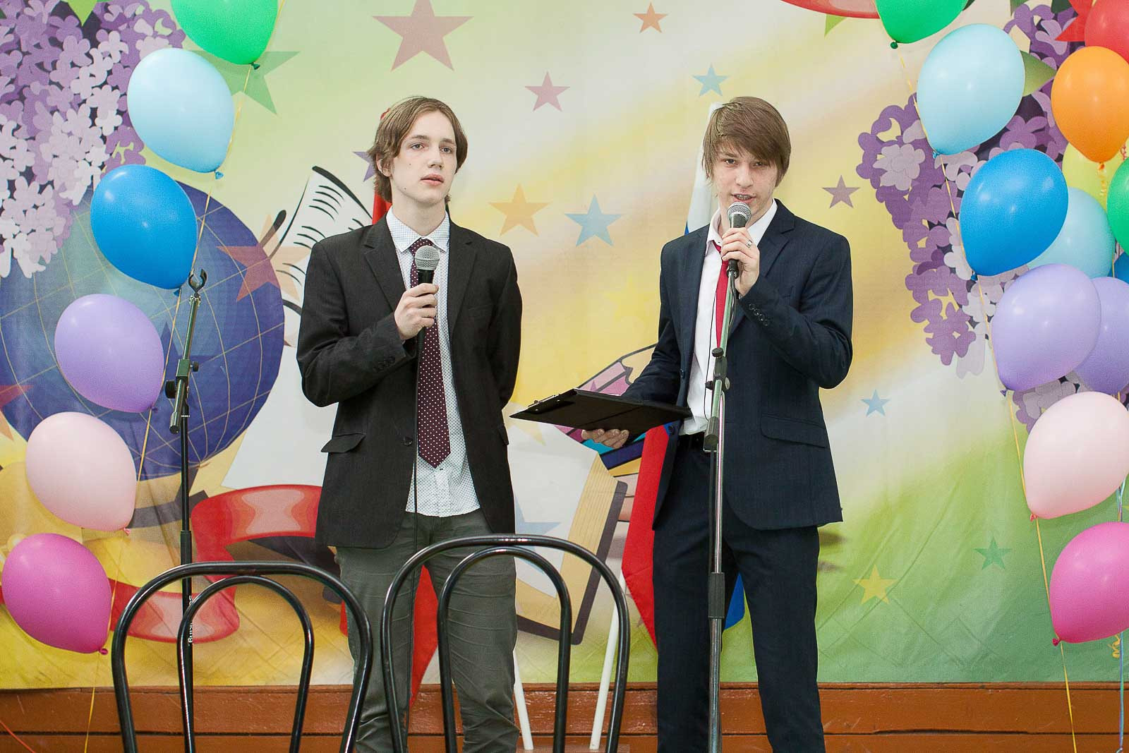 Два юноши в темных костюмах говорят в микрофон на фоне праздничных шаров.