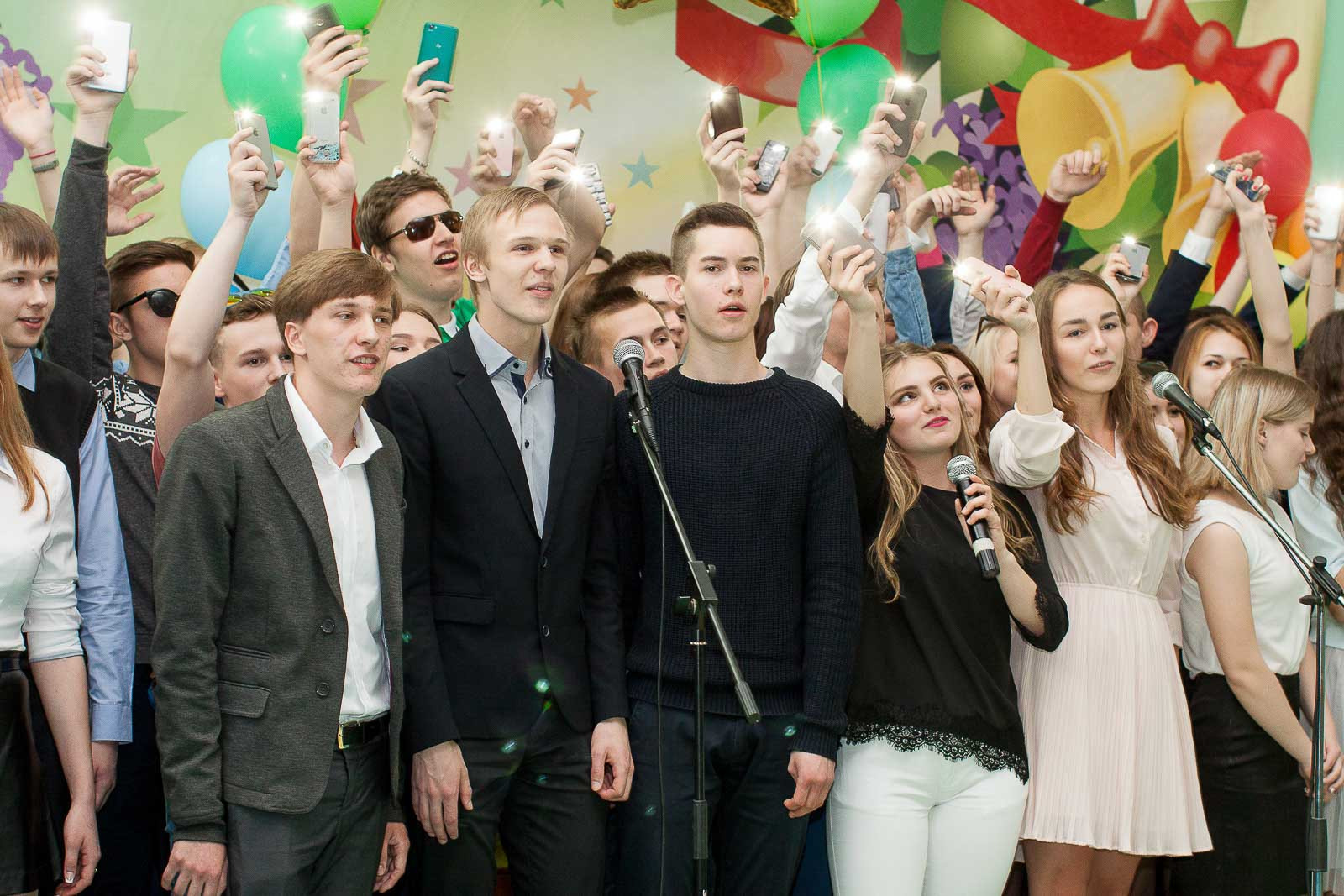 Выпускники с телефонами в руках поют песню в микрофоны.