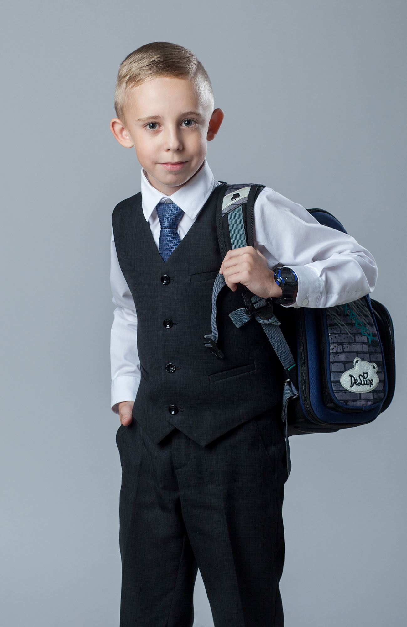 Поколенный студийный портрет выпускника 4 класса в тройке без пиджака на сером фоне с портфелем на левом плече.