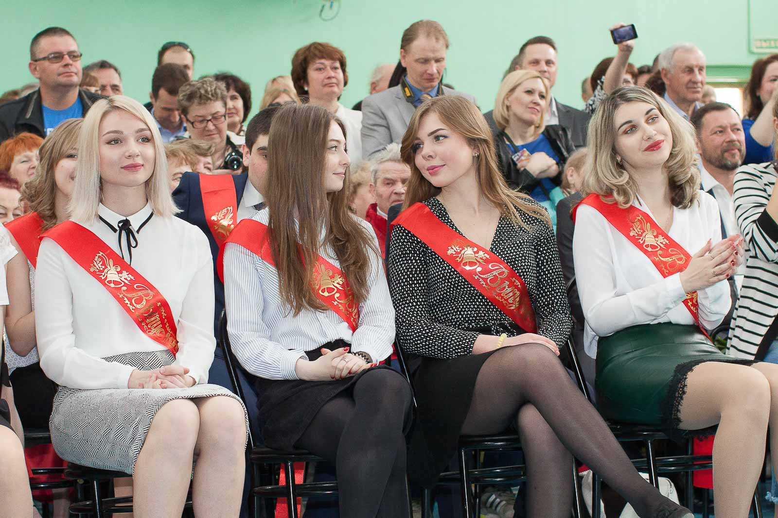 Выпускницы 11 класса сидят на стульях на фоне зрителей-родителей.