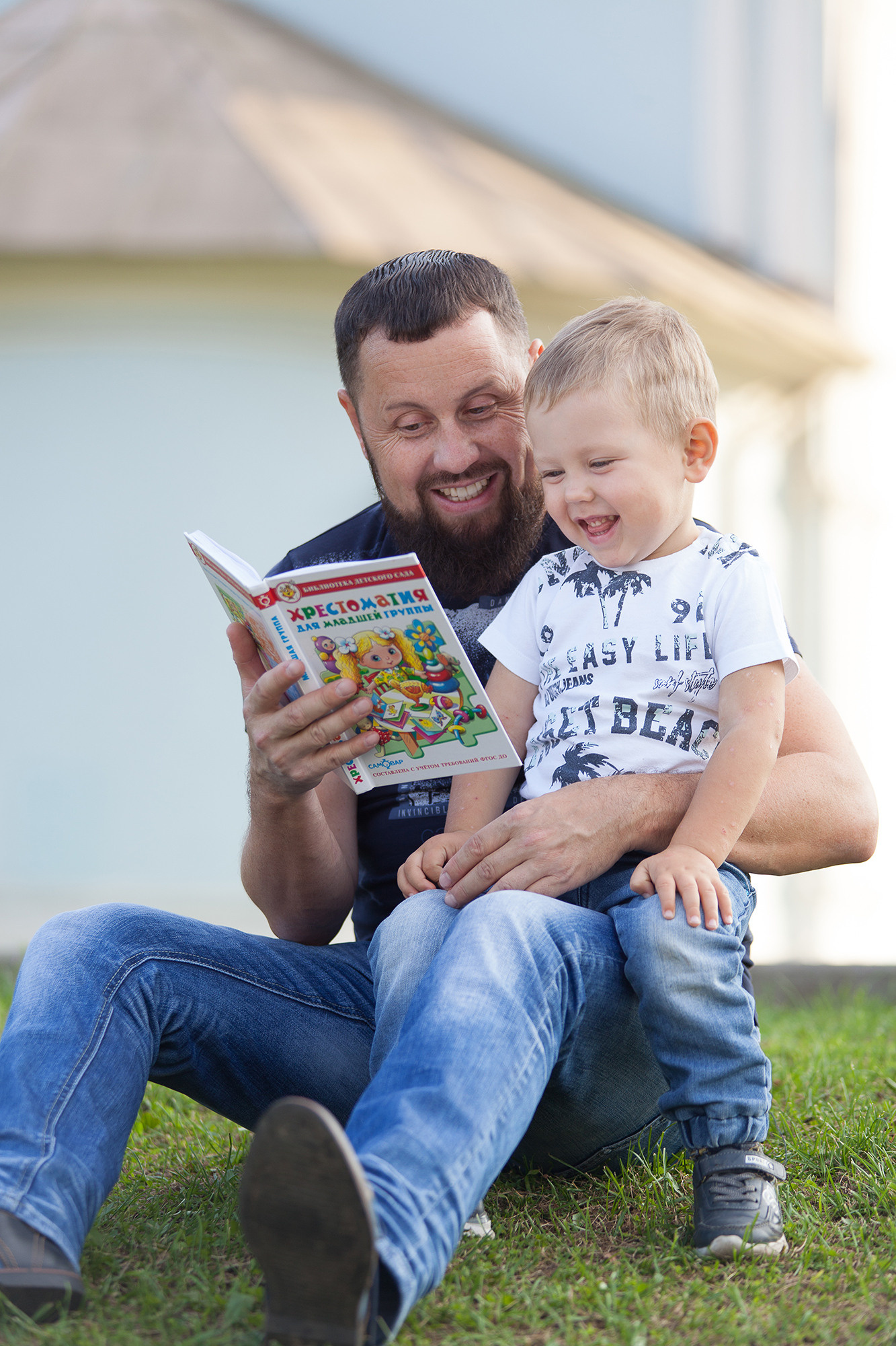 Мальчик в белой футболке и джинсах сидит на колене у папы и рассматривает книгу.