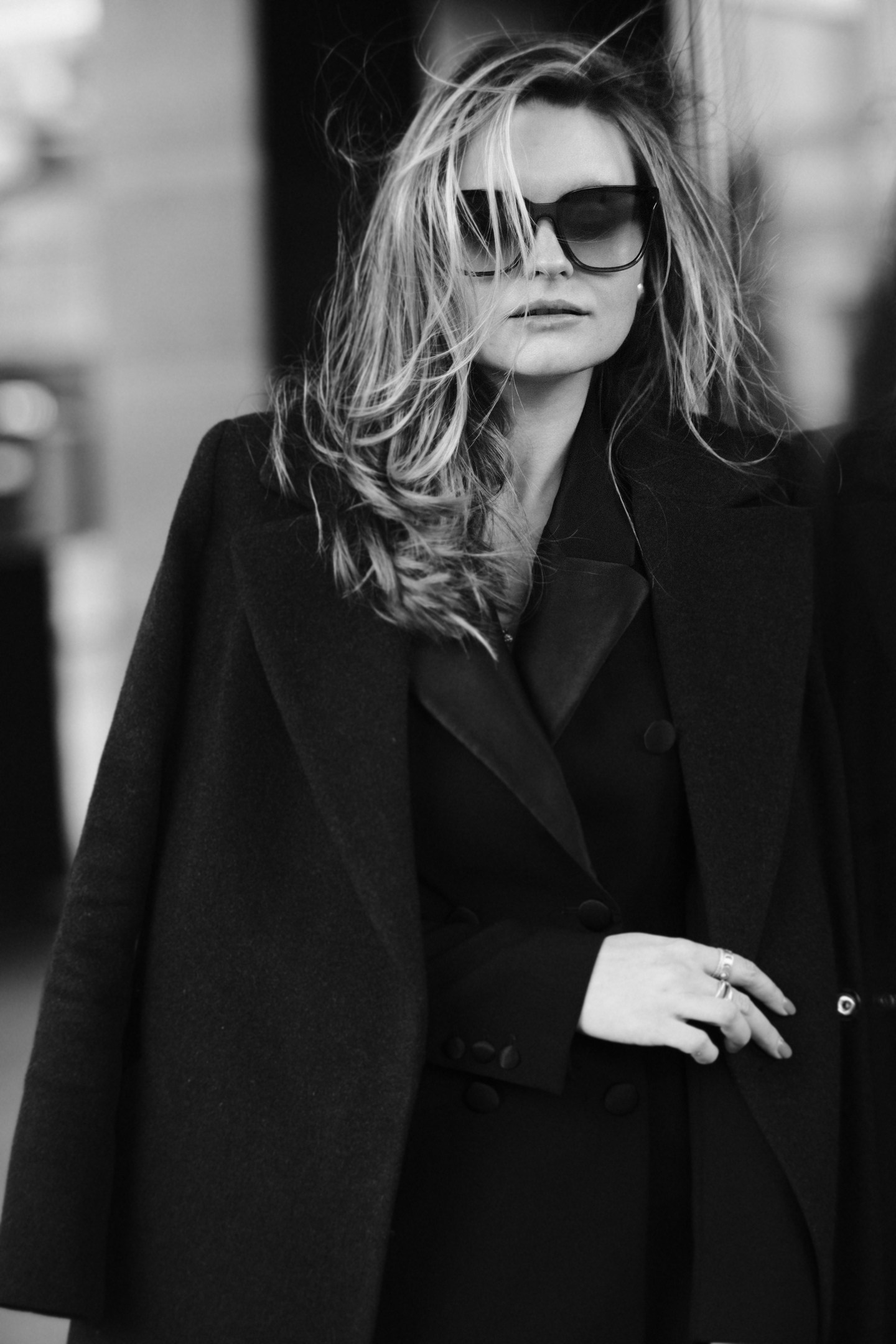 Черно-белый уличный портрет девушки в черном пальто-кейпе и солнцезащитных очках, развевающиеся волосы, модный образ