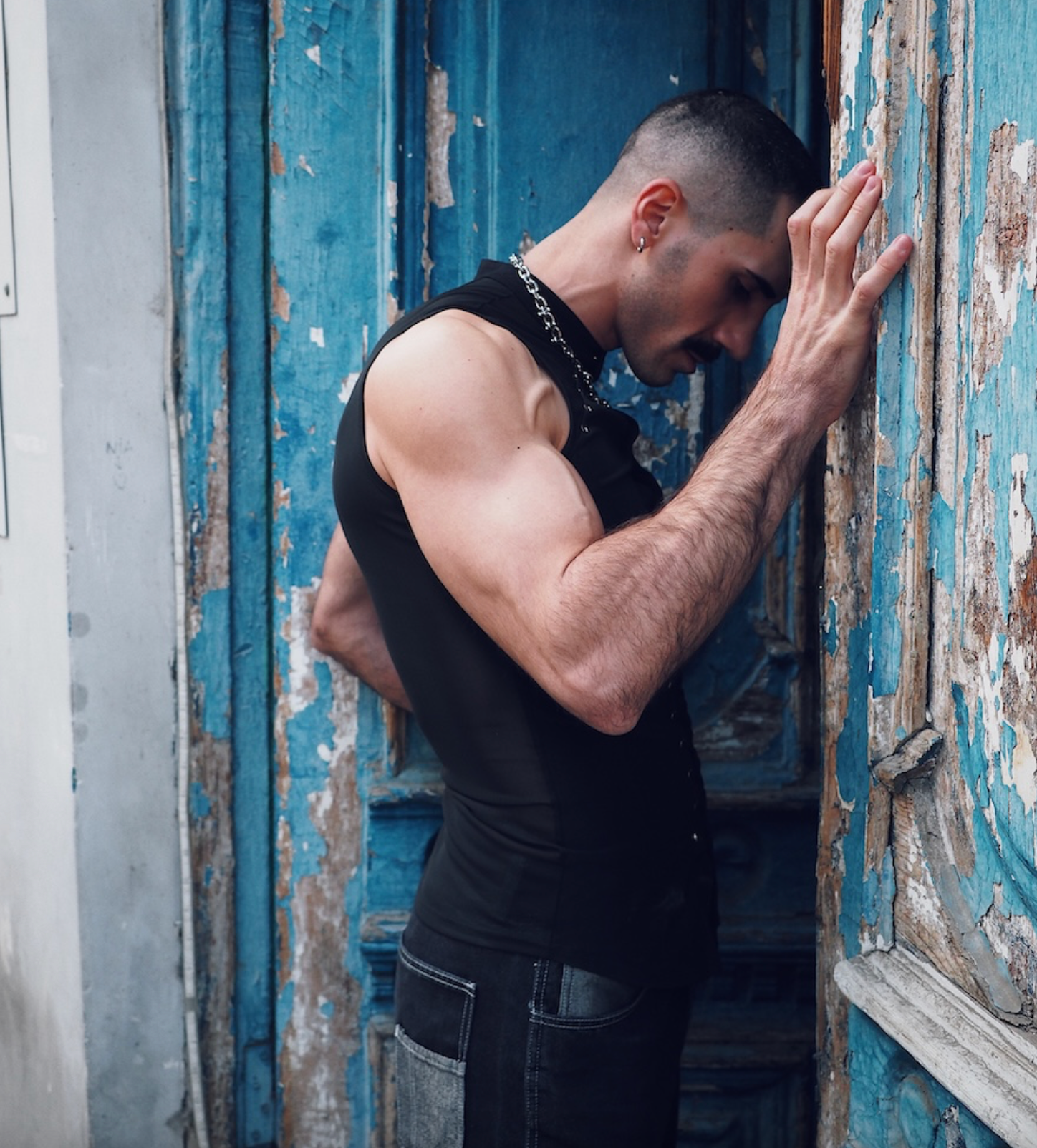 Male fashion portrait by Sergio MAiiS featuring a muscular model in a black sleeveless top standing against a weathered blue wooden doorway. Editorial menswear photography with urban aesthetics, dramatic mood, and strong masculine presence.
