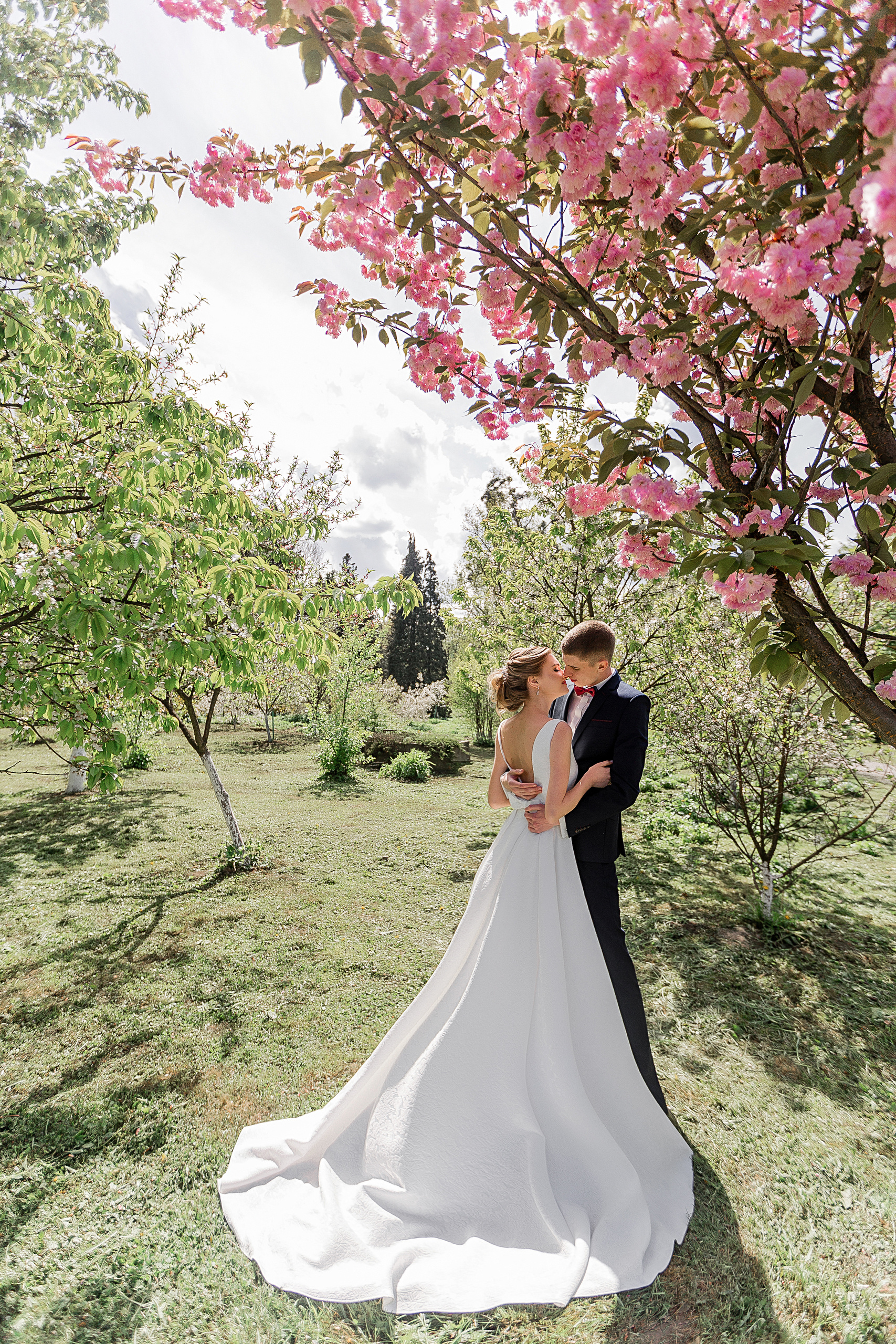 Нежная невеста обнимает стильного жениха в весеннем саду сакуры