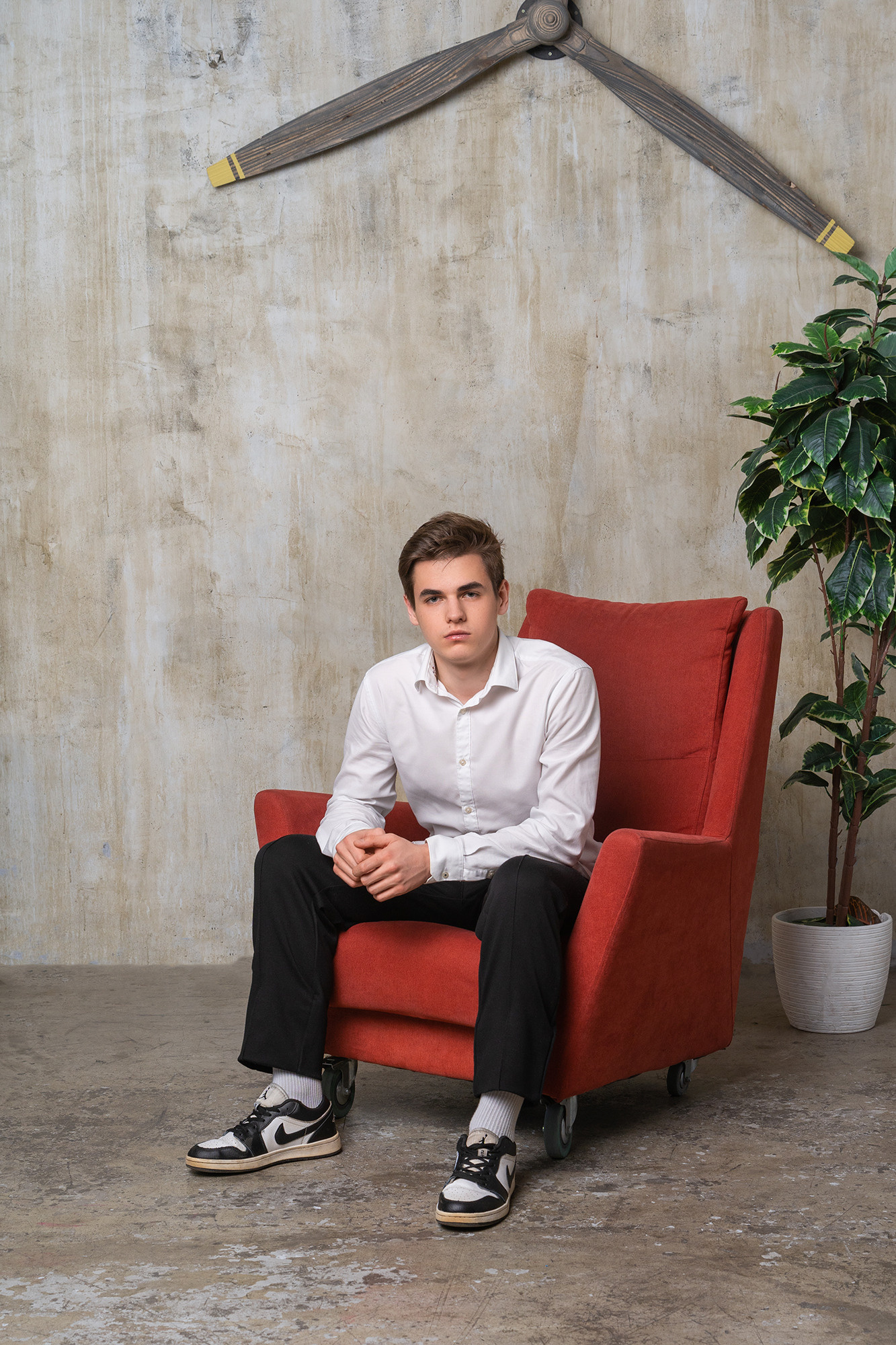 Юноша в белой рубашке, черных брюках и кроссовках сидит в красном кресле.