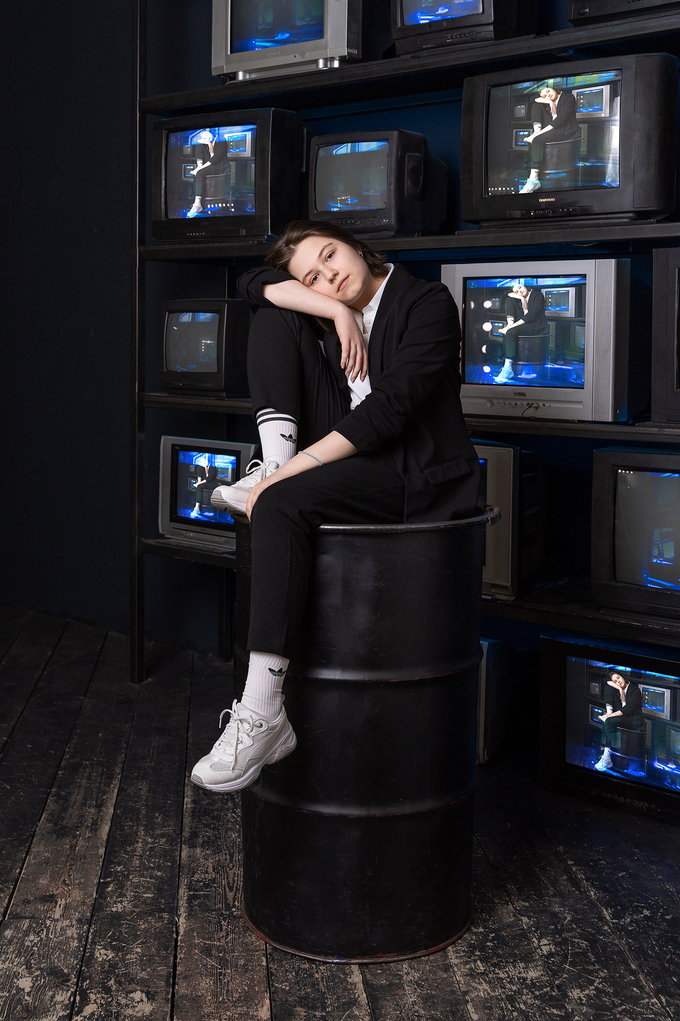 Девушка сидит на черной железной бочке на фоне телевизоров.