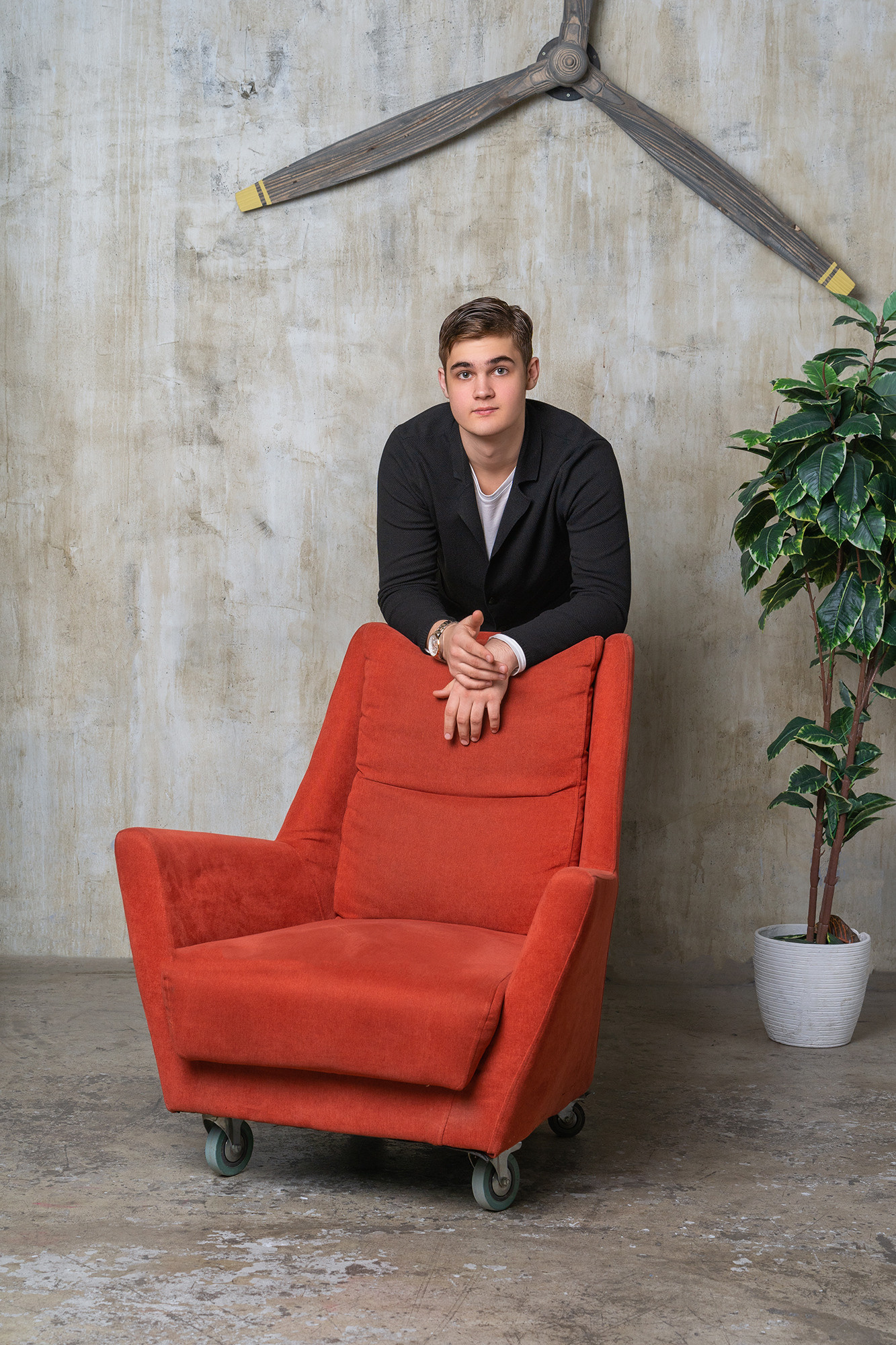 Юноша стоит, облокотившись на спинку красного кресла.