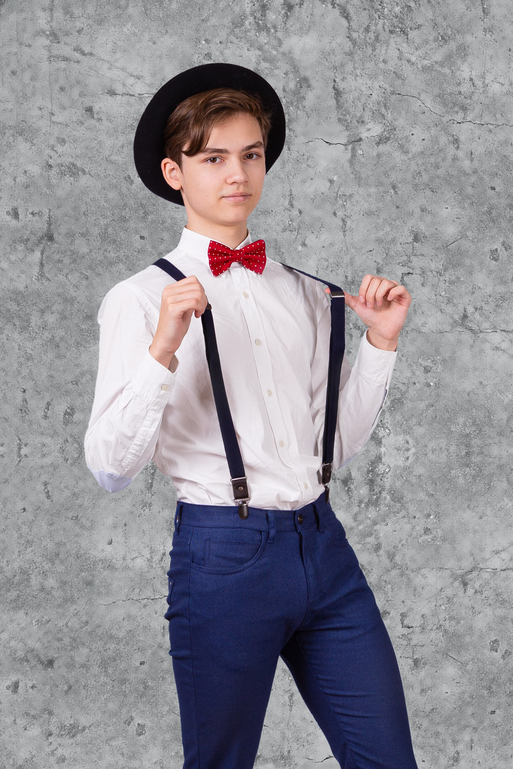 Юноша в черной шляпе, красном галстуке-бабочке и синих брюках с подтяжками.