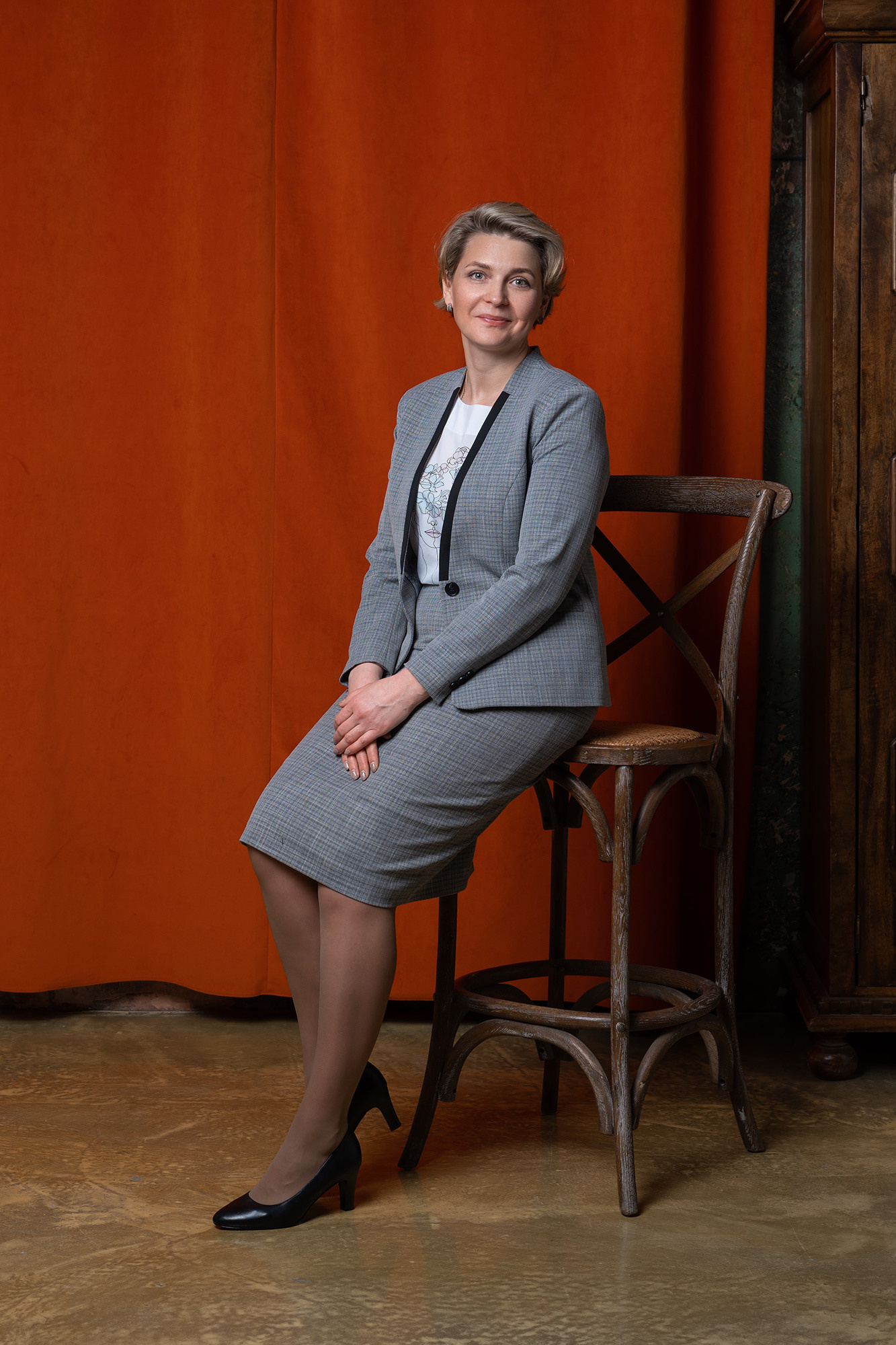 Женщина в сером костюме сидит на высоком деревянном стуле на фоне красной занавеси.