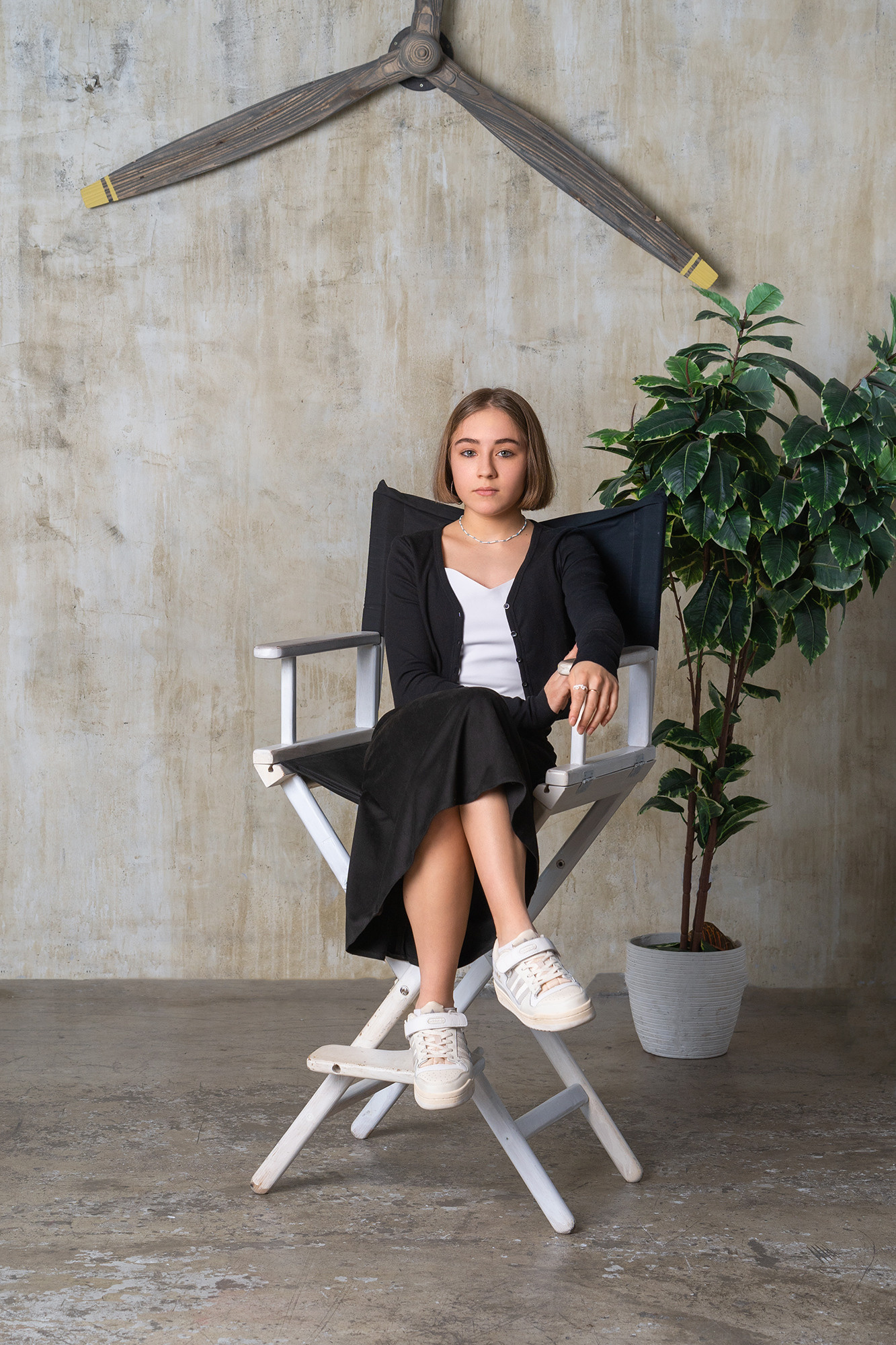 Девушка сидит на высоком режиссерском стуле. Сзади высокое растение в белом горшке.
