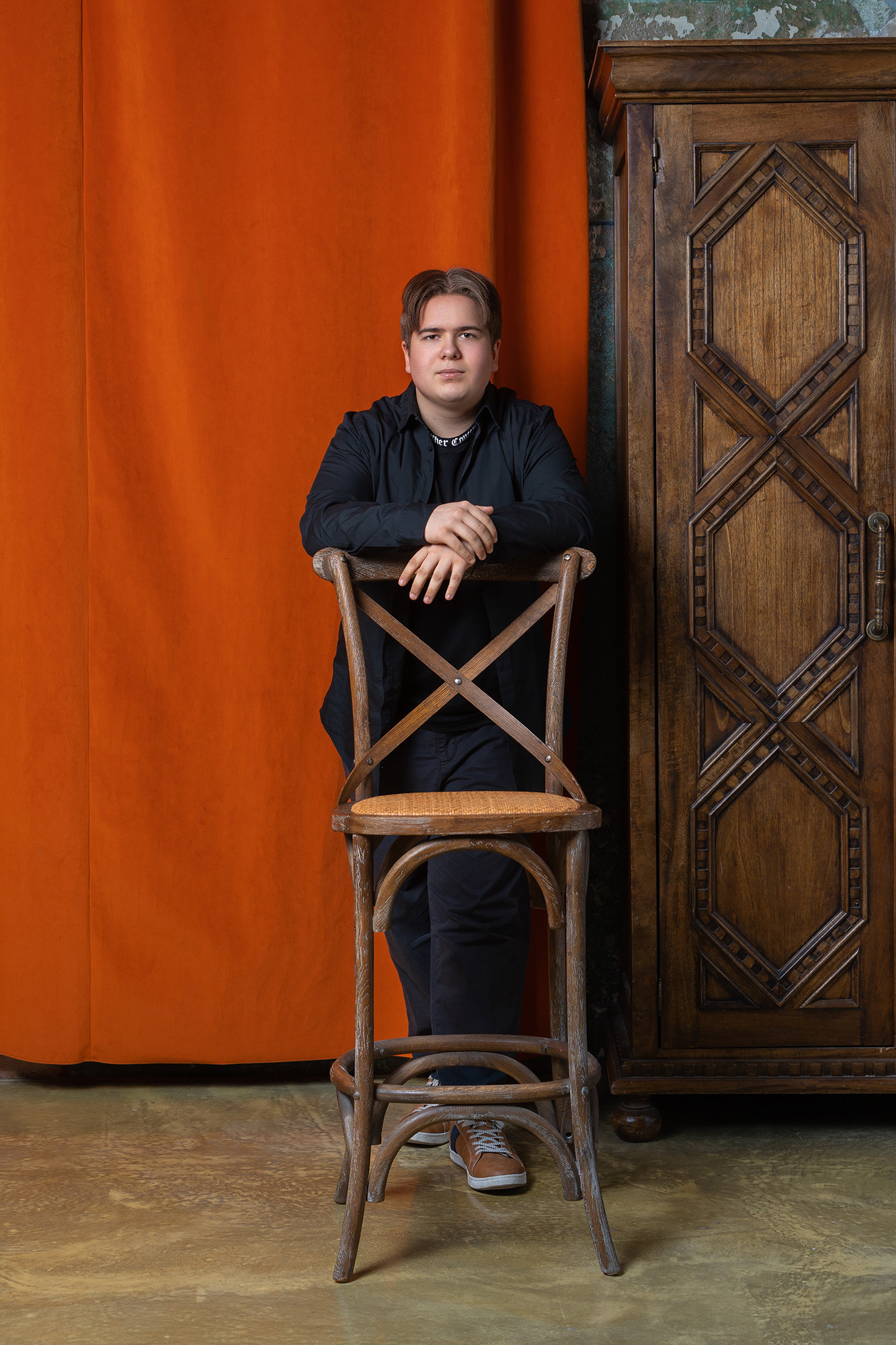 Юноша стоит на фоне красного занавеса и деревянного шкафа. На переднем плане высокий деревянный стул.