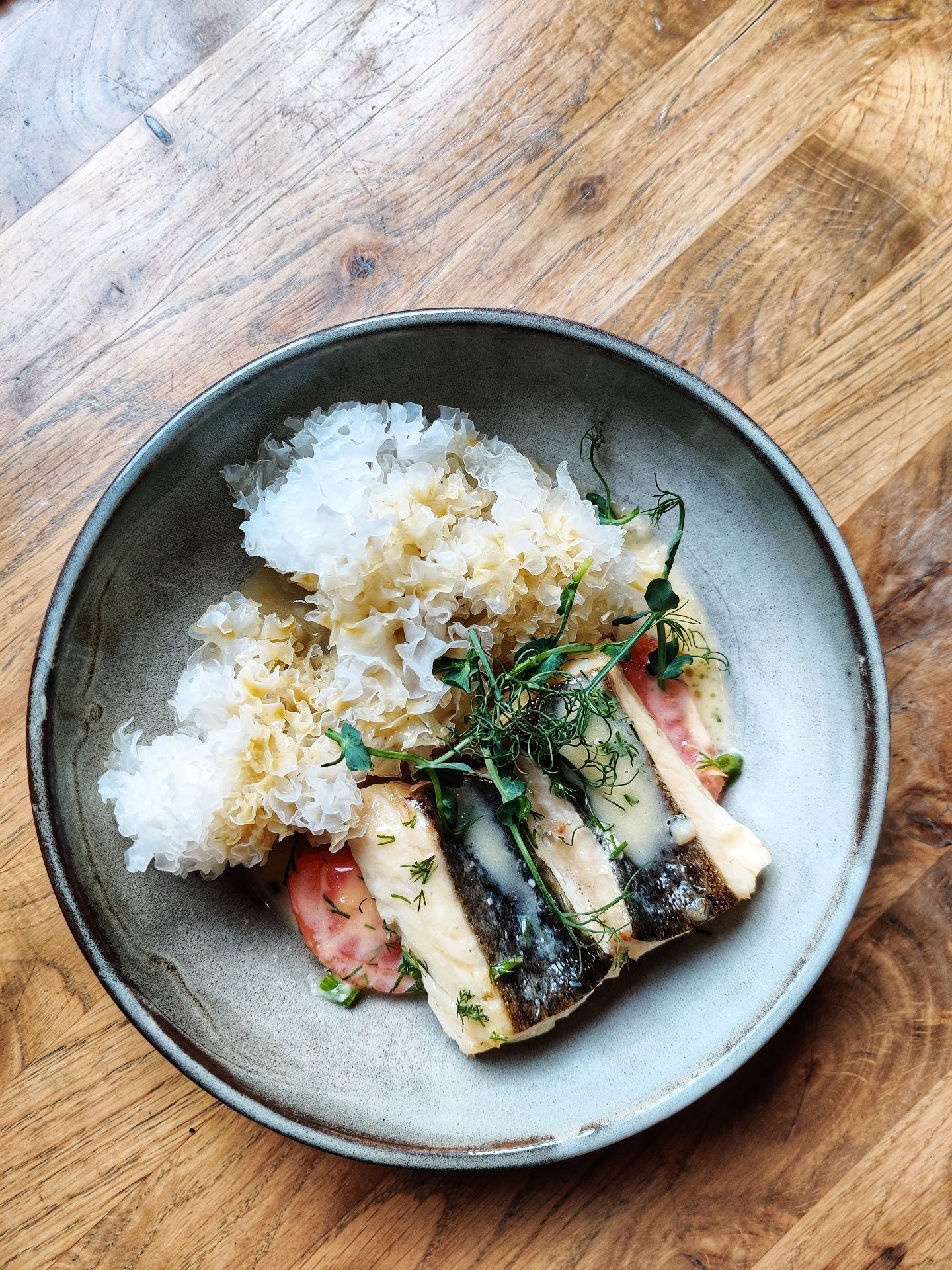 Блюдо с кусочками рыбы, белыми грибами-тремелла, помидорами и зеленью, поданное на серой тарелке на деревянном столе