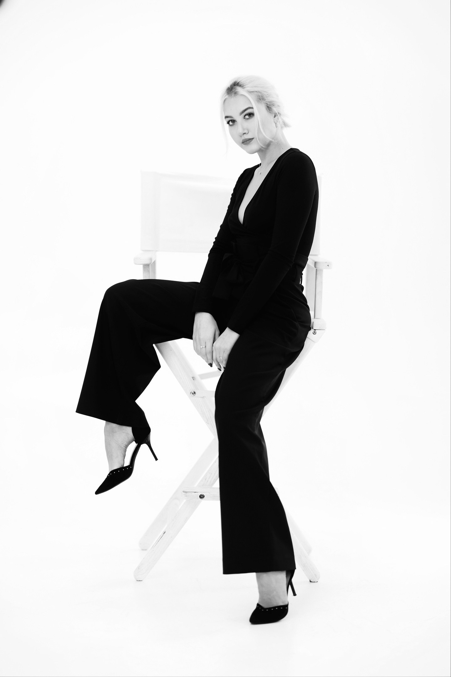 Черно-белый студийный портрет девушки в черном костюме на белом режиссерском кресле, полный рост, классическая поза