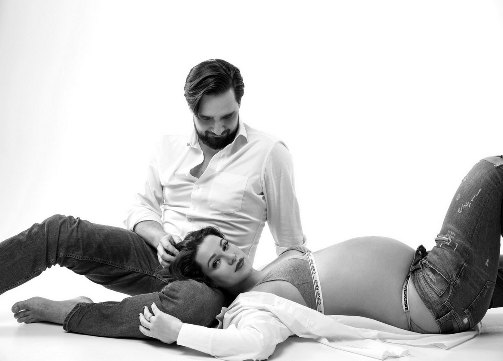 Беременная пара расположилась на белом полу мужчина нежно гладит женщину по голове.