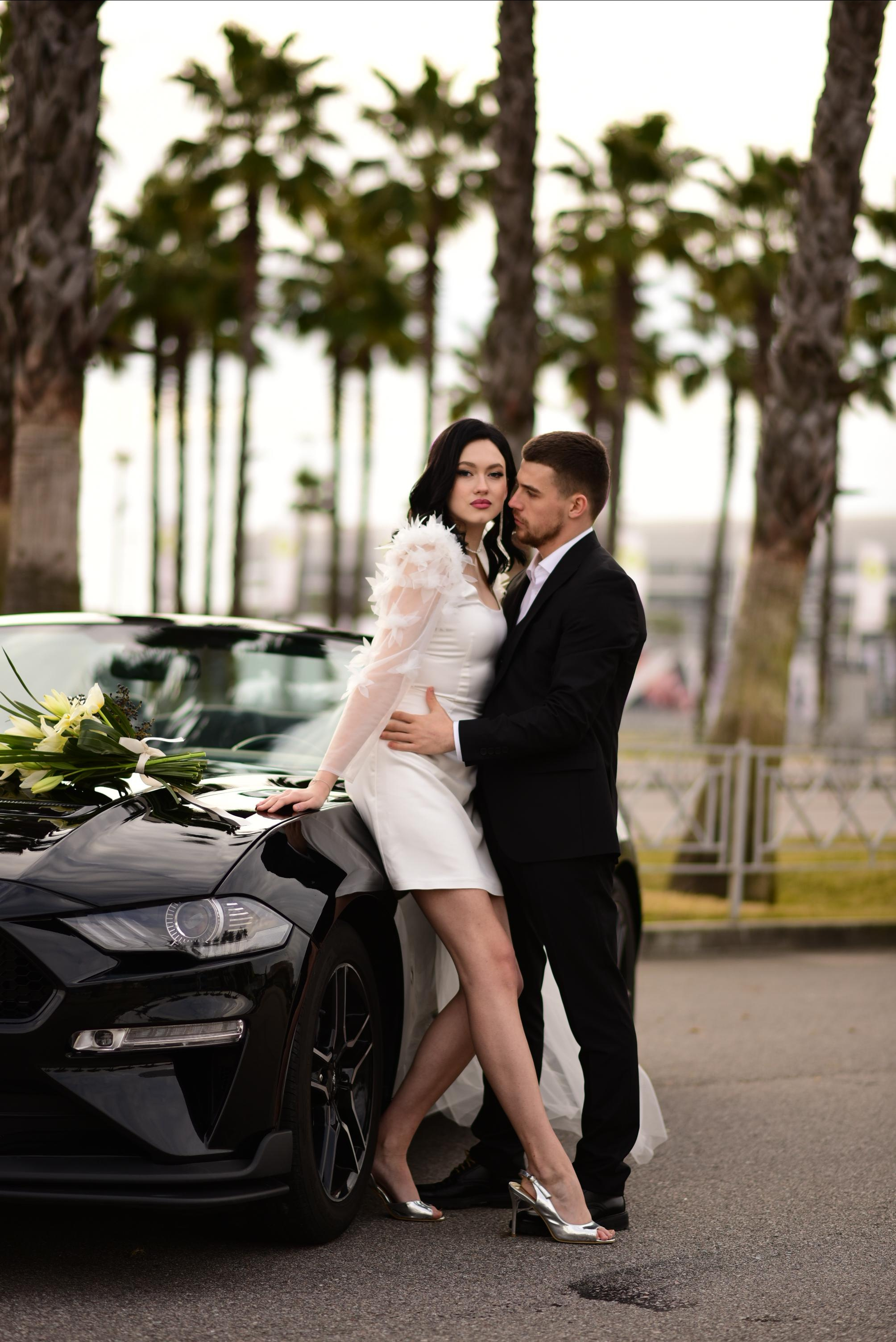 Жених и невеста с букетом в руках на фоне черного кабриолета.